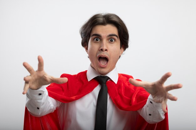 Ragazzo giovane supereroe sorpreso che guarda l'obbiettivo che indossa la cravatta tendendo le mani alla macchina fotografica isolata su priorità bassa bianca