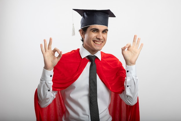 Ragazzo giovane supereroe sorridente che indossa cravatta e cappello laureato che mostra gesto giusto isolato su priorità bassa bianca