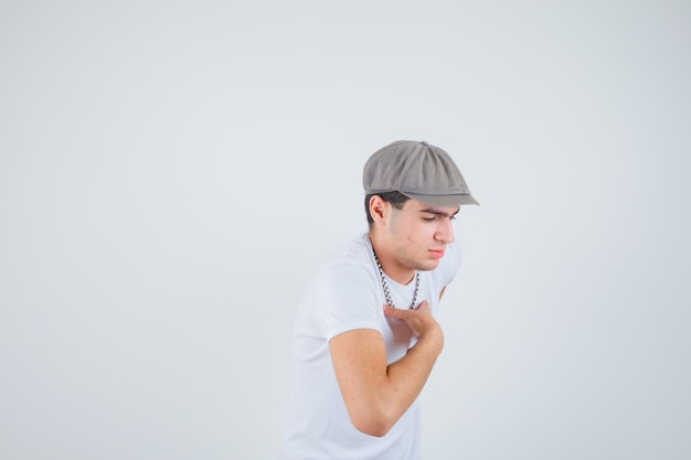 Ragazzo giovane tenendo la mano sul petto in t-shirt, cappello e guardando pensieroso. vista frontale.