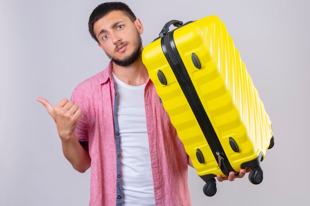 Ragazzo giovane viaggiatore bello che tiene la valigia che punta con il dito al lato che guarda l'obbiettivo con l'espressione confusa sul viso in piedi su sfondo bianco