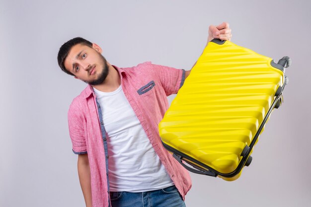 Ragazzo giovane viaggiatore bello che tiene la valigia che sembra confuso con l'espressione triste sul viso in piedi su sfondo bianco