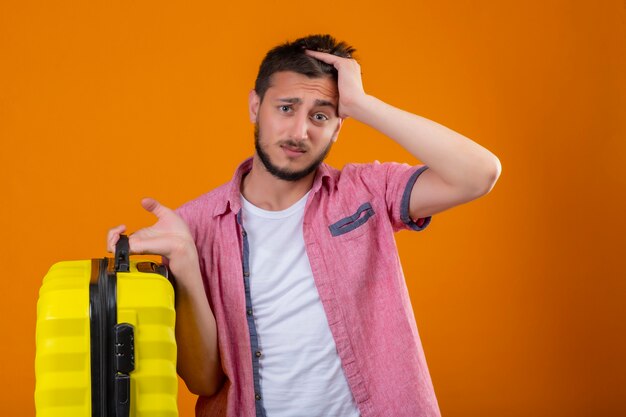 Ragazzo giovane viaggiatore bello che tiene la valigia in piedi con la mano sulla testa per errore ricorda il concetto di cattiva memoria di errore su sfondo arancione