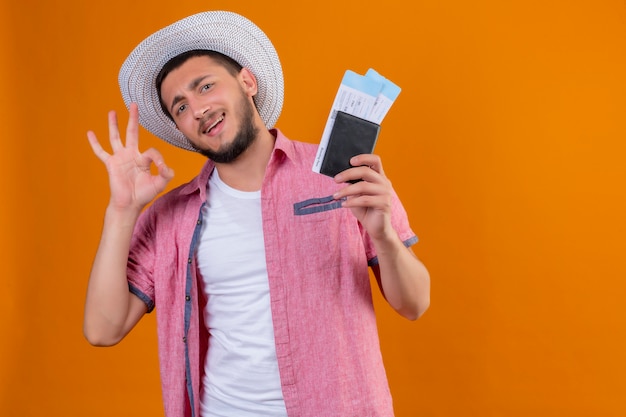 Ragazzo giovane viaggiatore bello con cappello estivo in possesso di biglietti aerei guardando la fotocamera uscita e felice sorridente allegramente facendo segno ok pronto a viaggiare in piedi su sfondo arancione