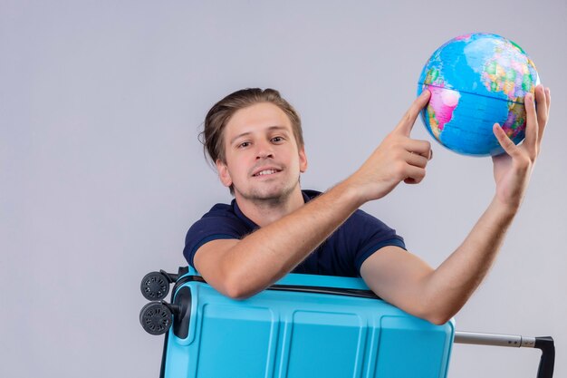 Ragazzo giovane viaggiatore bello in piedi con la valigia tenendo il globo guardando la fotocamera con espressione fiduciosa sorridente su sfondo bianco