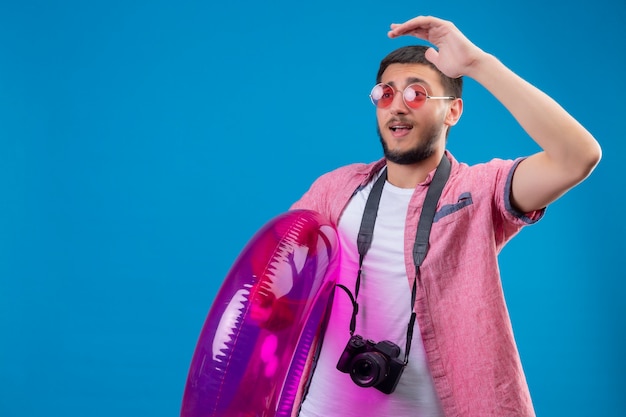 Ragazzo giovane viaggiatore bello indossando occhiali da sole in piedi con anello gonfiabile che osserva da parte agitando con mano felice e positivo su sfondo blu