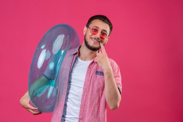 Ragazzo giovane viaggiatore bello indossando occhiali da sole tenendo anello gonfiabile guardando la telecamera sorridendo allegramente indicando con il dito alla bocca in piedi su sfondo rosa