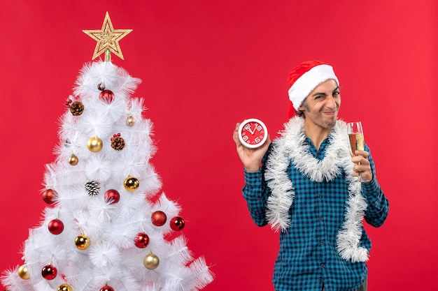 Ragazzo giovane viso aspro con cappello di Babbo Natale e alzando un bicchiere di vino e tenendo l'orologio in piedi vicino all'albero di Natale sul rosso