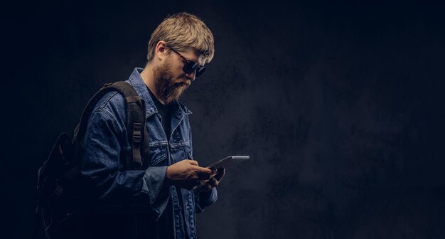 Ragazzo hipster barbuto che indossa occhiali da sole e zaino vestito con una giacca di jeans utilizzando un tablet PC. Isolato su uno sfondo scuro.