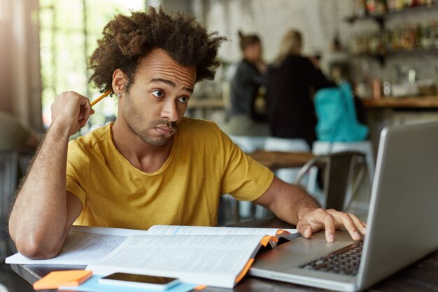 Ragazzo hipster con i capelli folti seduto alla mensa universitaria grattandosi la testa con la matita cercando di capire come svolgere un compito difficile utilizzando internet per aiutare