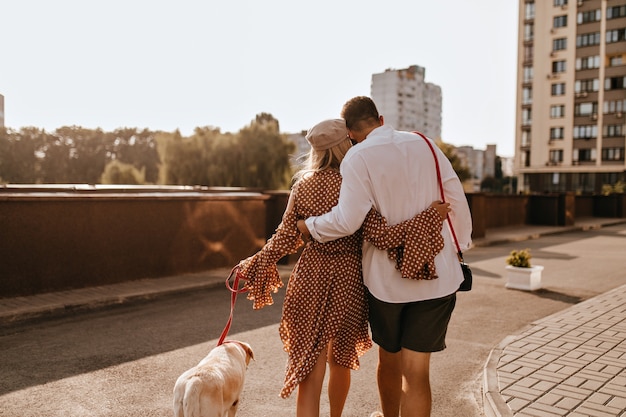 Ragazzo in camicia bianca e pantaloncini sta abbracciando la sua ragazza in abito a pois. Coppie che camminano il loro labrador bianco.