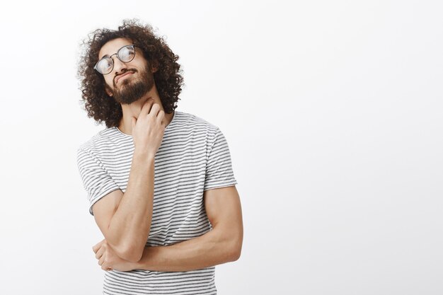 ragazzo ispanico attraente confuso incerto con elegante taglio di capelli ricci in occhiali, testa inclinata e grattarsi il collo, pensare, essere interrogato