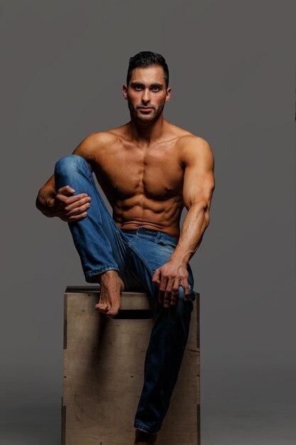Ragazzo muscoloso attraente senza camicia in blue jeans seduto sul podio su sfondo grigio