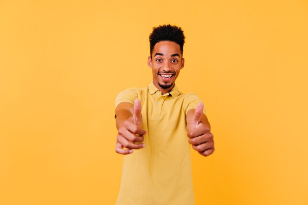 Ragazzo nero raffinato che mostra i pollici in su con un sorriso sorpreso. Foto dell'interno dell'uomo africano di buon umore con l'acconciatura divertente.