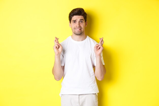 Ragazzo nervoso incrociando le dita per buona fortuna, sperando in qualcosa, in piedi su sfondo giallo