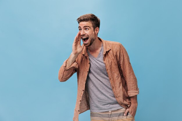 Ragazzo positivo dagli occhi grigi con barba fresca in camicia alla moda e pantaloni beige a strisce che guardano lontano e urlano sul muro blu