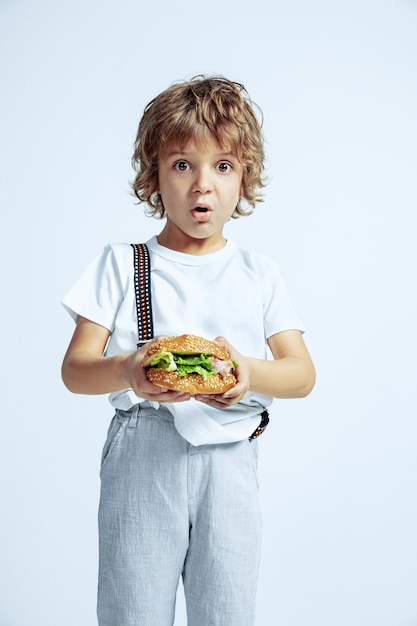 Ragazzo riccio abbastanza giovane in abbigliamento casual sulla parete bianca. Mangiare hamburger. Bambino in età prescolare maschio caucasico con emozioni facciali luminose. Infanzia, espressione, divertimento, fast food. stupito.