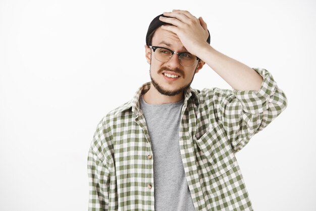 Ragazzo sconvolto e tormentato con barba e baffi in berretto nero da hipster e occhiali che tiene la mano sulla testa che si acciglia e si acciglia in una situazione problematica e imbarazzante