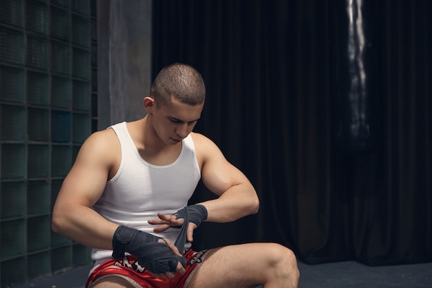 Ragazzo serio con la testa rasata e bicipiti forti che ha concentrato l'espressione mentre indossa bende da boxe nere al polso prima di allenarsi in palestra. Persone, sport, arti marziali e kickboxing