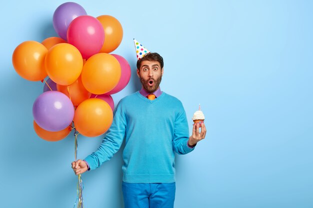 Ragazzo sorpreso con cappello di compleanno e palloncini in posa in maglione blu