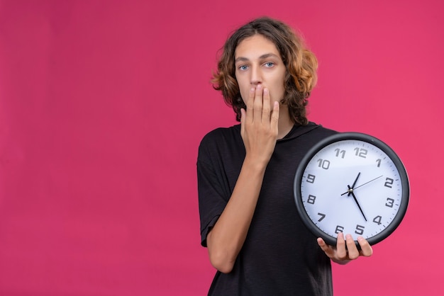 Ragazzo sorpreso con i capelli lunghi in maglietta nera che tiene un orologio da parete e si coprì la bocca con la mano su sfondo rosa