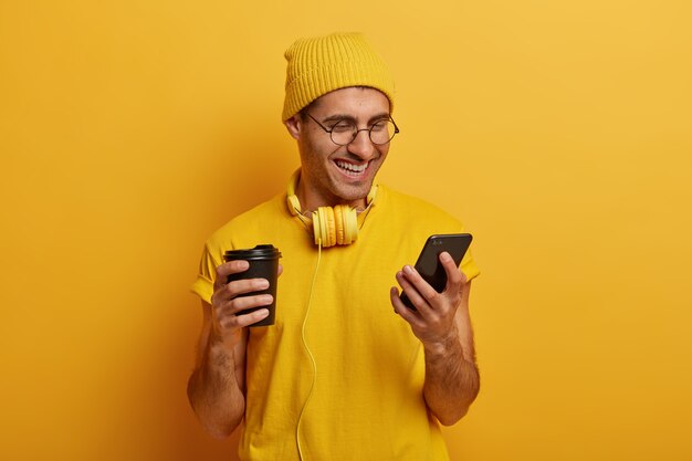 Ragazzo sorridente allegro guarda un video divertente tramite smartphone, beve gustose bevande calde dal bicchiere di carta, indossa una maglietta e un cappello gialli