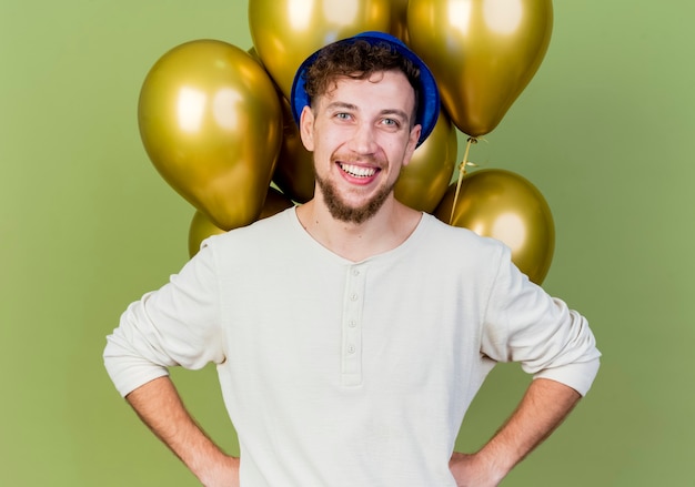 Ragazzo sorridente bello partito slavo che indossa il cappello del partito in piedi davanti a palloncini che guarda l'obbiettivo tenendo le mani sulla vita isolato su sfondo verde oliva