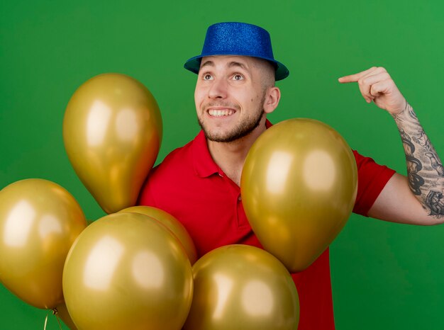 Ragazzo sorridente bello partito slavo che indossa il cappello del partito in piedi dietro i palloncini guardando il lato che punta a palloncini isolati su priorità bassa verde