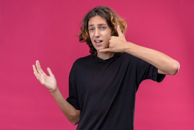 Ragazzo sorridente con capelli lunghi in maglietta nera che fa chiamata con la sua mano sulla parete rosa