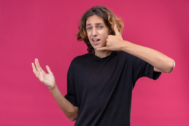 Ragazzo sorridente con capelli lunghi in maglietta nera che fa chiamata con la sua mano sulla parete rosa