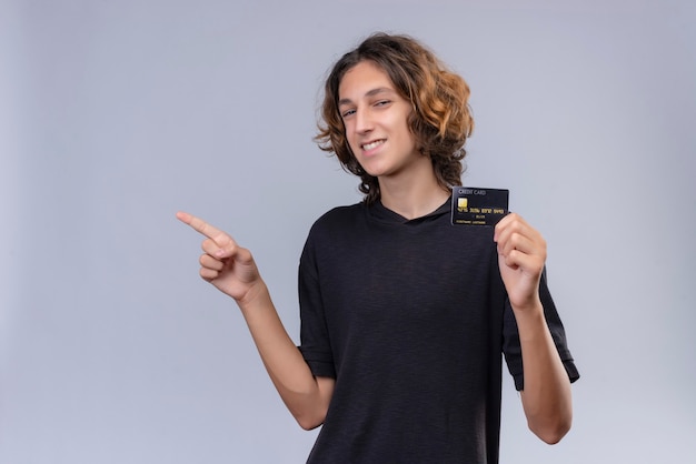 Ragazzo sorridente con capelli lunghi in maglietta nera che tiene una carta di credito e punta a lato sul muro bianco