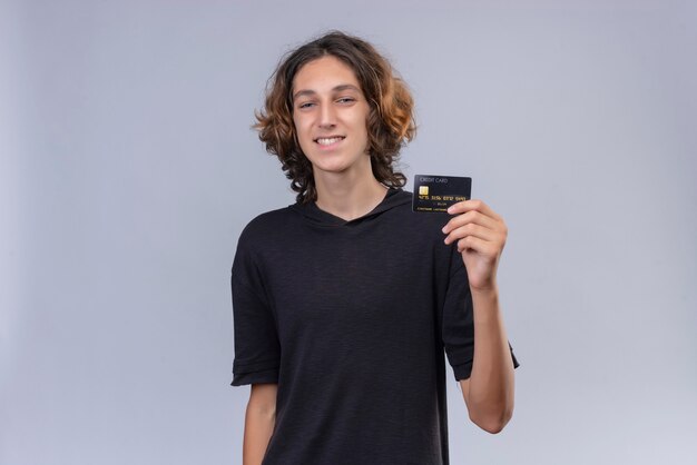 Ragazzo sorridente con capelli lunghi in maglietta nera che tiene una carta di credito sul muro bianco