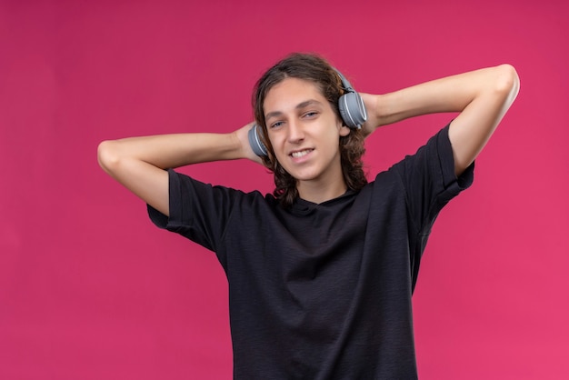 Ragazzo sorridente con i capelli lunghi in maglietta nera ascolta musica dalle cuffie e ha afferrato la testa sul muro rosa