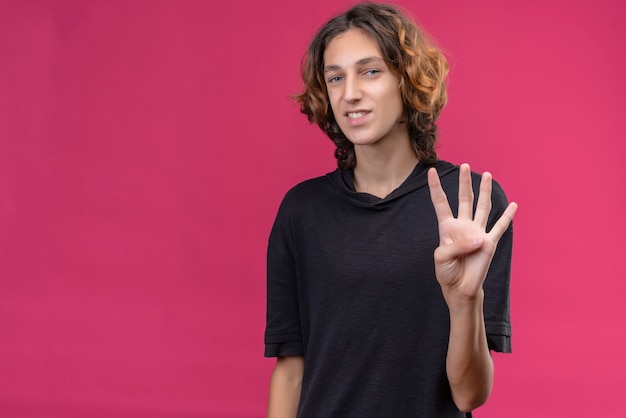 Ragazzo sorridente con i capelli lunghi in maglietta nera che mostra quattro con le dita sul muro rosa