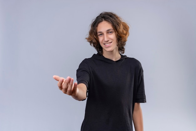 Ragazzo sorridente con i capelli lunghi in maglietta nera tese la mano sul muro bianco