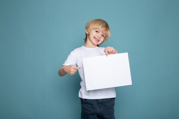 Ragazzo sorridente del bambino in maglietta e blue jeans bianche con pezzo di carta vuoto sul blu