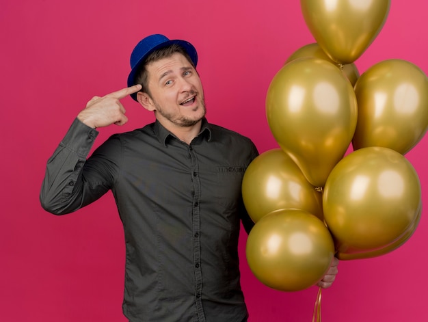 Ragazzo sorridente del partito giovane che indossa il cappello blu che tiene palloncini e punti a se stesso isolato sul rosa