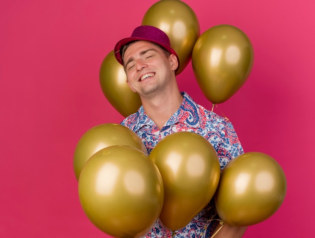 Ragazzo sorridente del partito giovane con gli occhi chiusi che indossa il cappello rosa in piedi tra palloncini isolati sul rosa