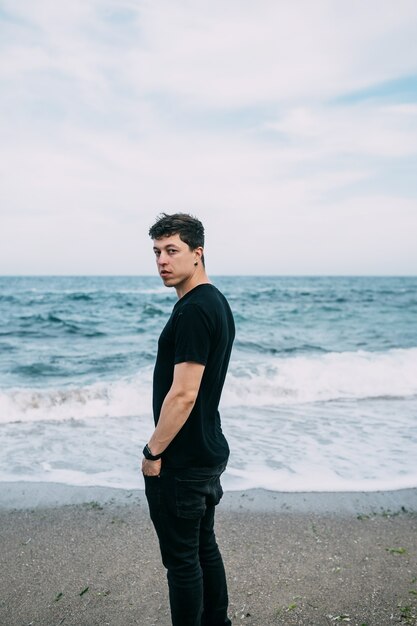 Ragazzo sorridente in una maglietta nera si trova sulla spiaggia sabbiosa.