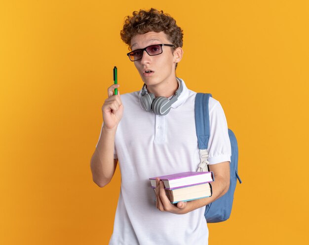 Ragazzo studente in abbigliamento casual con cuffie che indossa occhiali con zaino con libri e penna che sembra sorpreso di avere una nuova idea in piedi su sfondo arancione