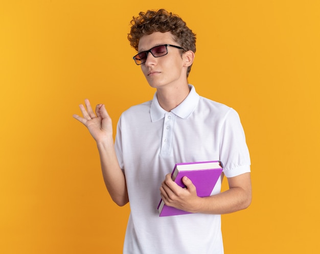 Ragazzo studente in abbigliamento casual con gli occhiali che tiene un libro guardando la telecamera felice e contento che mostra il segno ok in piedi su sfondo arancione