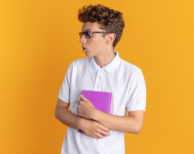 Ragazzo studente in abbigliamento casual con gli occhiali con in mano un libro che guarda da parte confuso