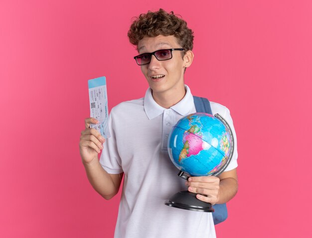 Ragazzo studente in abbigliamento casual con gli occhiali con lo zaino che tiene il globo e il biglietto aereo guardando la telecamera sorridendo allegramente in piedi su sfondo rosa