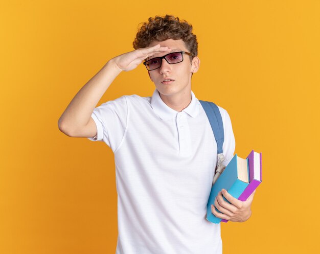 Ragazzo studente in abbigliamento casual con gli occhiali con zaino in mano libri che guarda lontano con la mano sopra la testa in cerca di qualcuno
