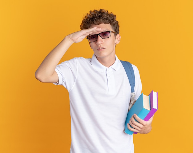 Ragazzo studente in abbigliamento casual con gli occhiali con zaino in mano libri che guarda lontano con la mano sopra la testa in cerca di qualcuno