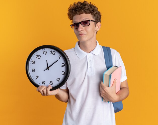 Ragazzo studente in abbigliamento casual con gli occhiali con zaino in mano libri e orologio da parete che guarda la telecamera con un sorriso fiducioso sul viso in piedi su sfondo arancione