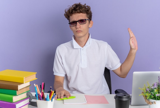 Ragazzo studente in polo bianca con gli occhiali seduto al tavolo con libri che guarda l'obbiettivo con faccia seria facendo gesto di difesa con la mano su sfondo blu
