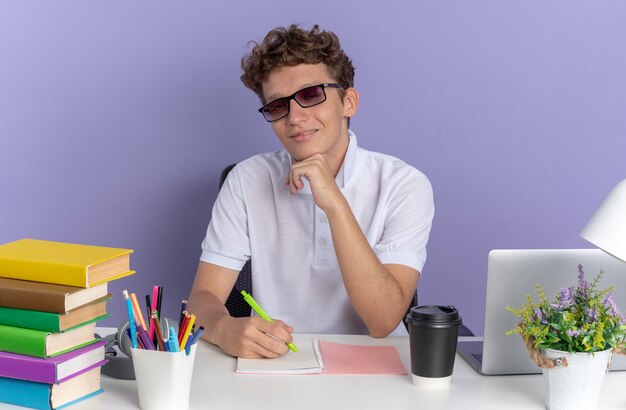 Ragazzo studente in polo bianca con gli occhiali seduto al tavolo con libri guardando la telecamera sorridendo fiducioso su sfondo blu