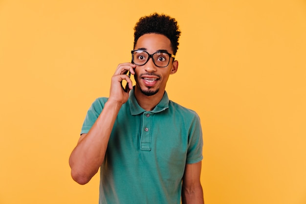 Ragazzo stupito in grandi bicchieri parlando al telefono. Ritratto dell'interno del ragazzo africano emotivo in maglietta verde che chiama qualcuno.