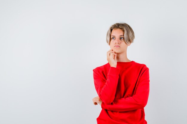 Ragazzo teenager che sta nella posa di pensiero in maglione rosso e che sembra preoccupato. vista frontale.