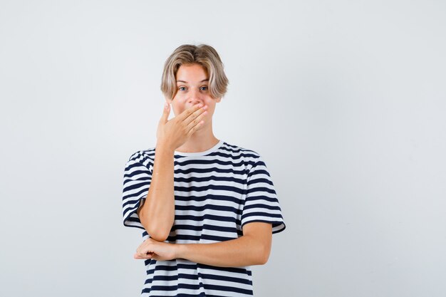 Ragazzo teenager che tiene la mano sulla bocca in maglietta e sembra stupito. vista frontale.